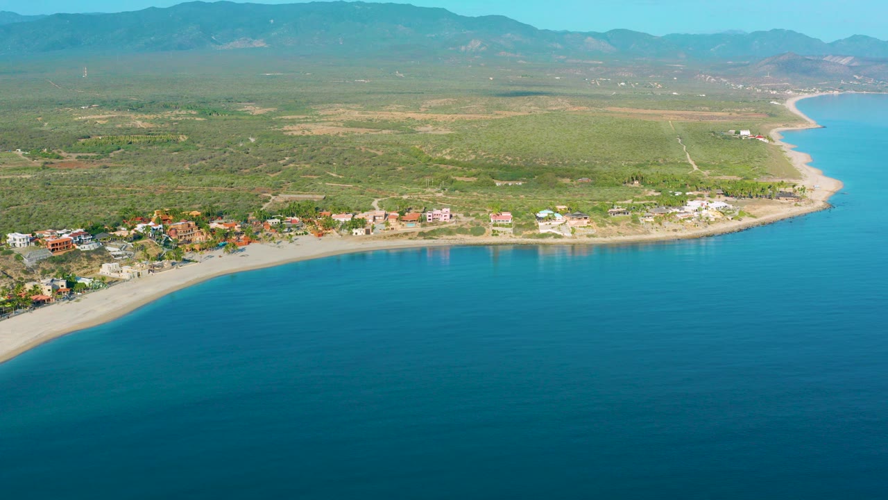Aerial view over La Ribera, Baja California Sur, with blue sea in the foreground, a curved shoreline with scattered buildings, green scrubland, distant mountains, and no visible people.