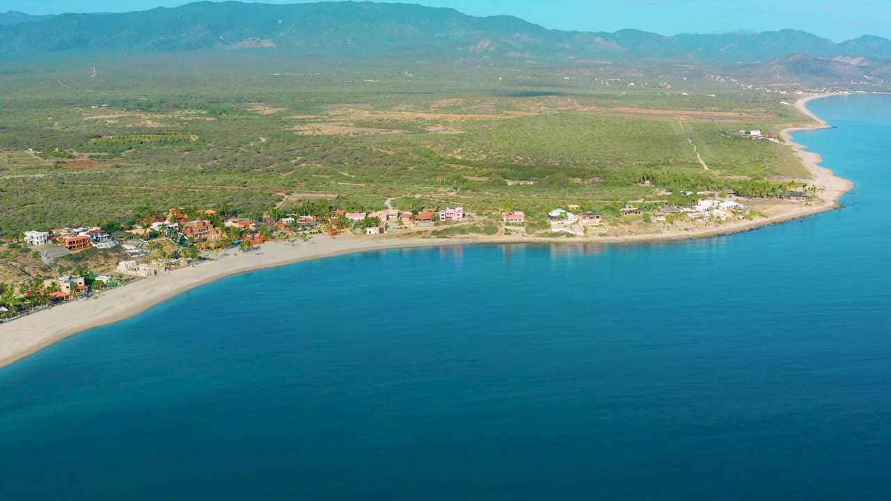 Drone-style view over La Ribera with deep blue sea, a pale beach curve, clustered shoreline buildings, open green inland terrain, distant mountains, and no visible people.