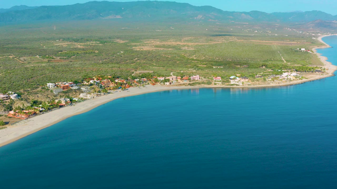 Opening frame of La Ribera, Baja California Sur from above, with turquoise water in front, a pale beach and small coastal buildings in the middle distance, mountains beyond, and no visible people.