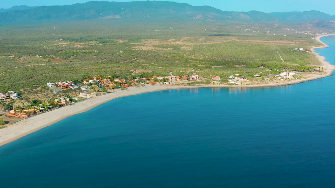 Aerial frame over La Ribera showing more open water in the foreground, a line of beach homes and low buildings along the sand, inland scrubland, distant mountains, and no visible people.