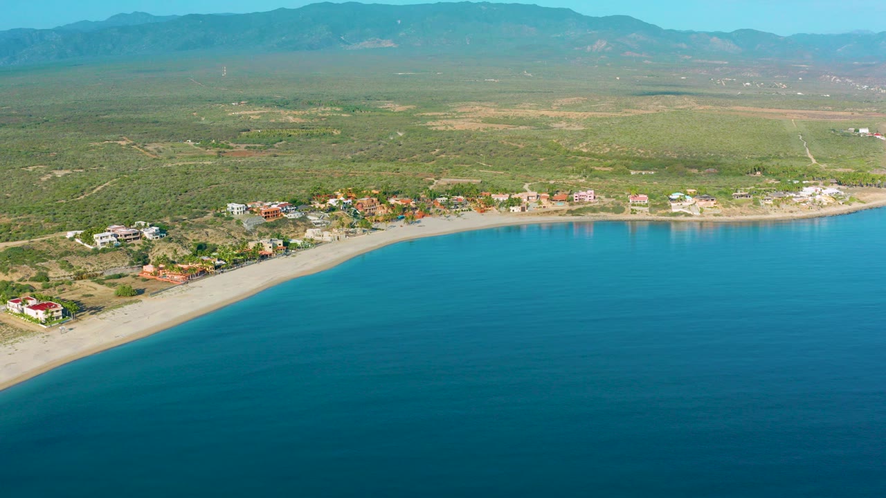 Opening aerial frame over La Ribera, Baja California Sur, with blue bay water in front, a curved pale beach, scattered coastal homes, inland scrubland, distant mountains, and no visible people.