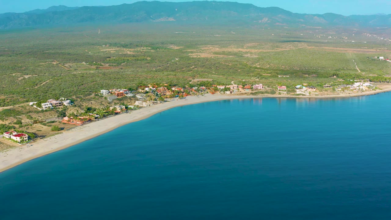 Aerial frame from La Ribera showing the shoreline slightly shifted across the view, with calm blue water below, low coastal buildings behind the sand, broad green terrain, mountains, and no visible people.