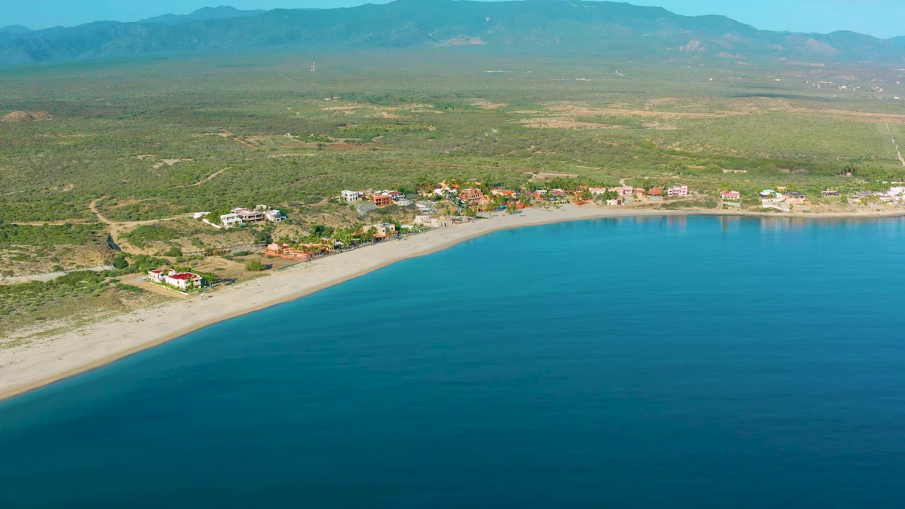 Drone view over La Ribera showing turquoise water along a pale beach, low coastal buildings just inland, broad green terrain, distant mountains, and no visible people.