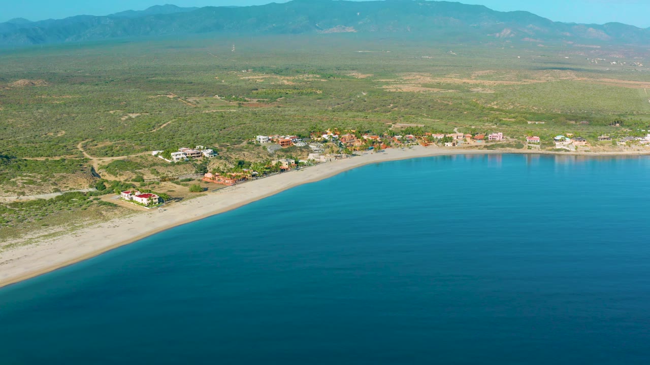 Wide aerial frame in La Ribera with deep blue water below, a sand shoreline, visible dirt tracks and homes inland, green scrubland, mountain ridges, and no visible people.