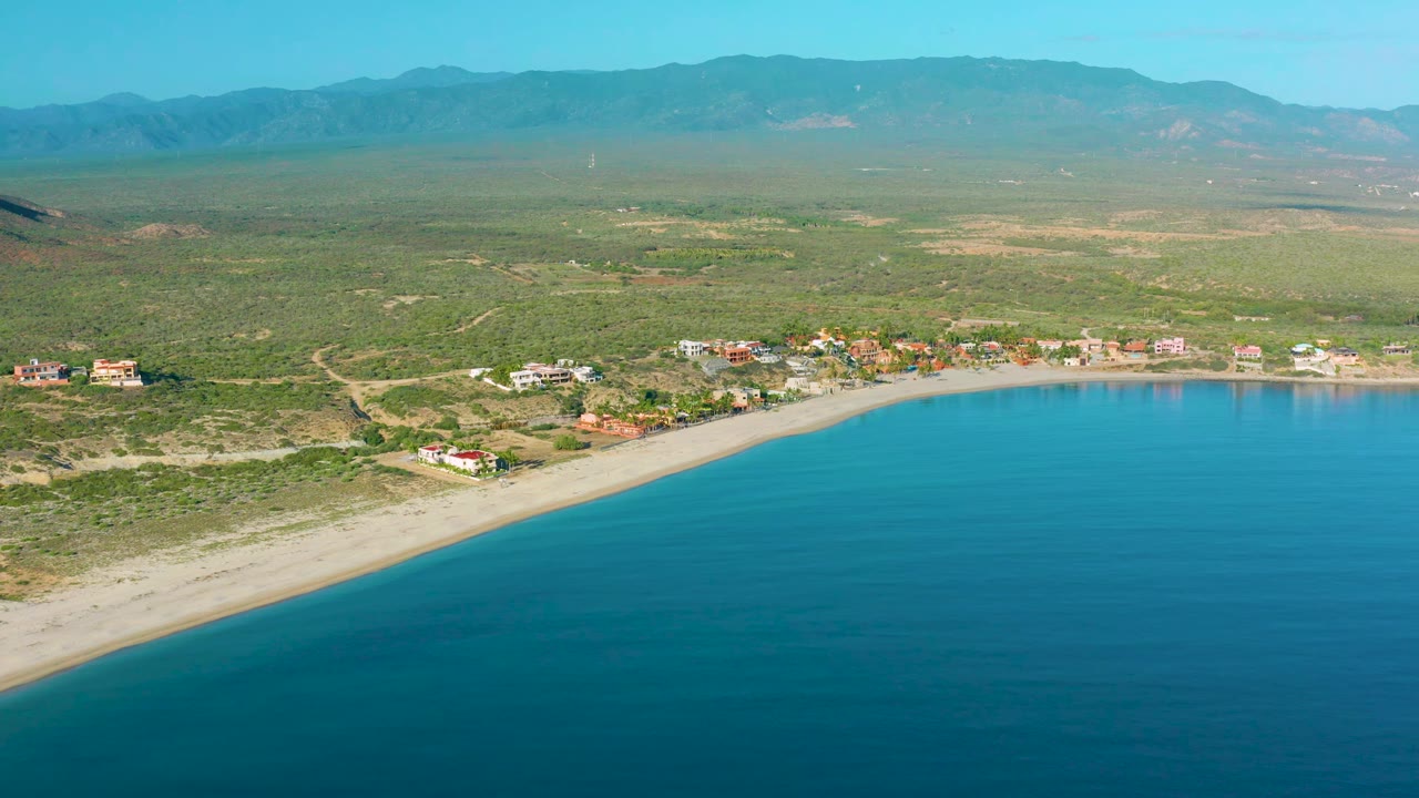 Drone view of curved shoreline, small houses, scrub vegetation, and hazy mountain horizon.