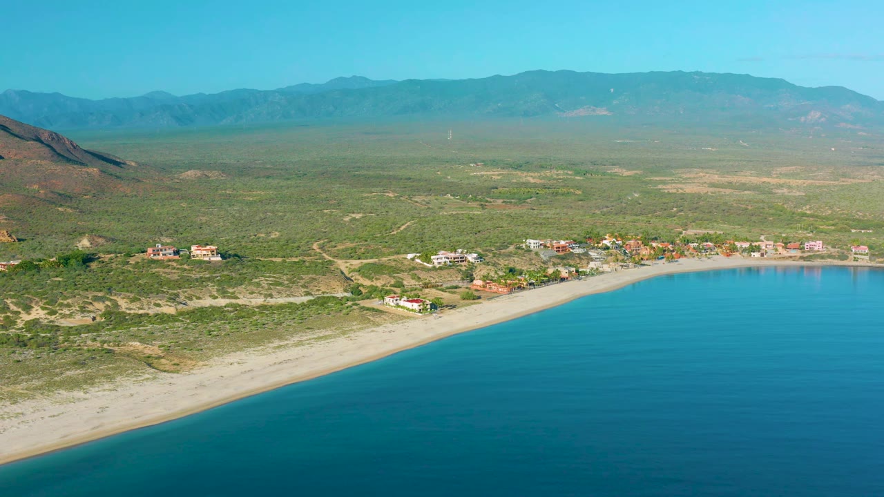Aerial coastline with turquoise water, pale beach, homes, green scrubland, and mountain ridge.