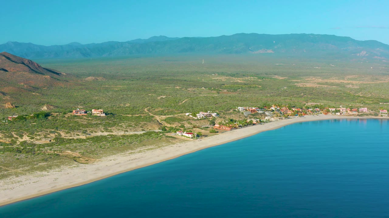 High aerial view over La Ribera with the curved bay at lower right, a thin sand beach, small coastal buildings, open scrubland, brown hills to the left, hazy mountain range, and no visible people.