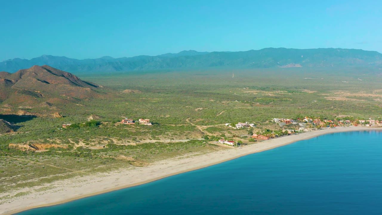 Wide aerial frame of La Ribera showing turquoise water, a narrow sandy shoreline, scattered coastal buildings, desert scrub, hills, distant ridges, and no visible people.