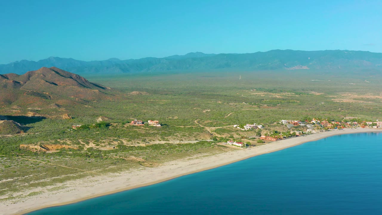 Aerial view over La Ribera with more sea filling the lower right, a bright beach line, small buildings near shore, green-brown scrubland, hills, mountains, and no visible people.