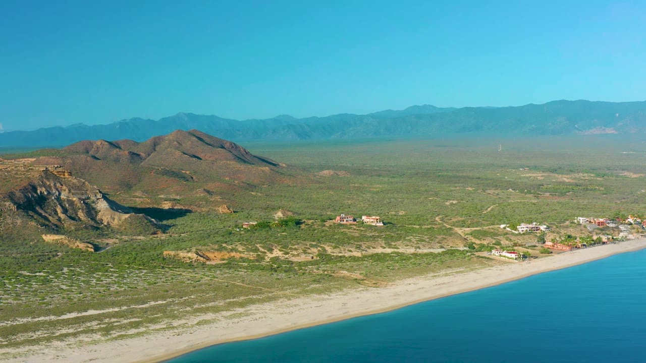 Wide aerial frame of La Ribera showing a calm blue shoreline curving past scrubland and small buildings, with rocky hills and distant ridges under clear sky, no visible people.
