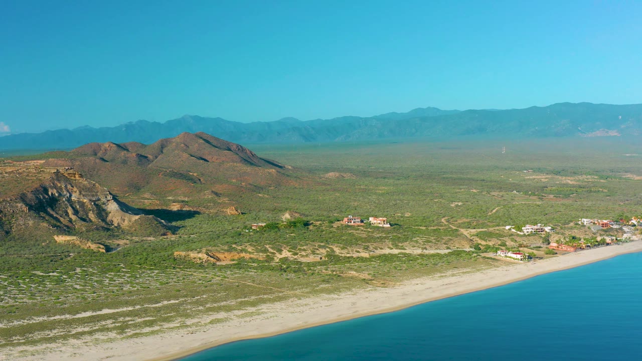 Drone-like view over La Ribera with turquoise water filling the lower right edge, a light sand beach, desert flats, clustered buildings, rocky hills, and hazy mountains, no visible people.