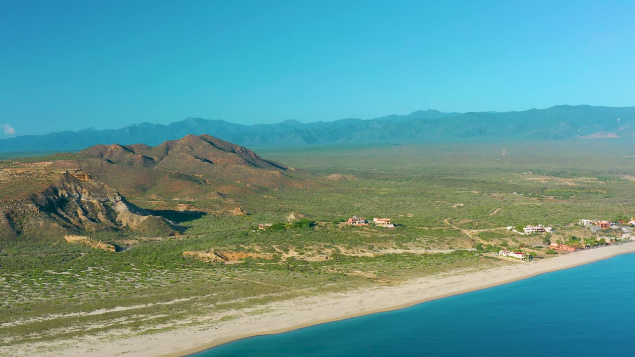 Aerial coastline with open sea, narrow beach, desert vegetation, scattered buildings, and rocky hills.