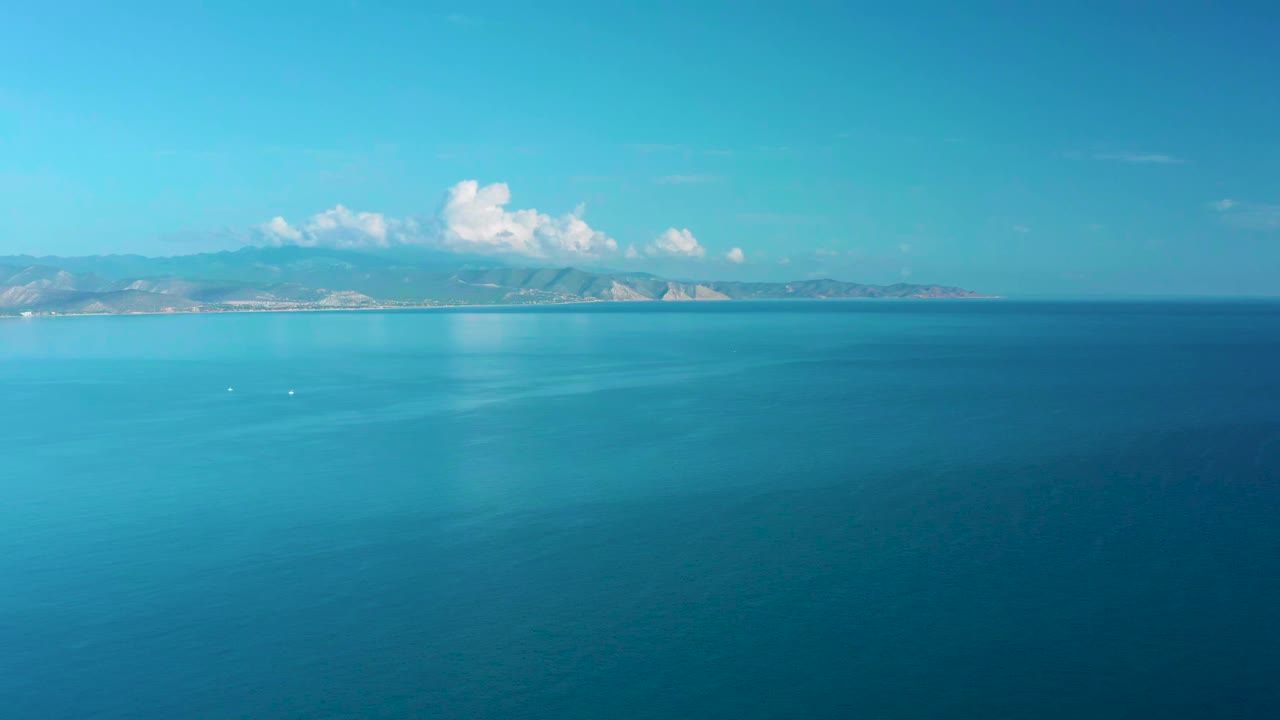 At 0:01, the La Ribera aerial view holds on blue water with faint surface texture, low mountains in the distance, scattered white clouds, and no visible people.