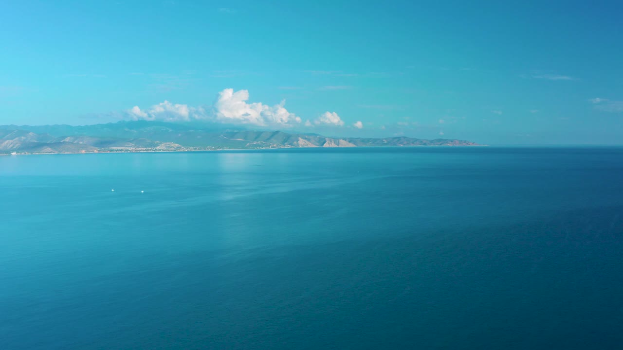 At 0:03, open ocean dominates the foreground while distant coastal mountains remain below a bright sky with small white clouds, with no visible people.