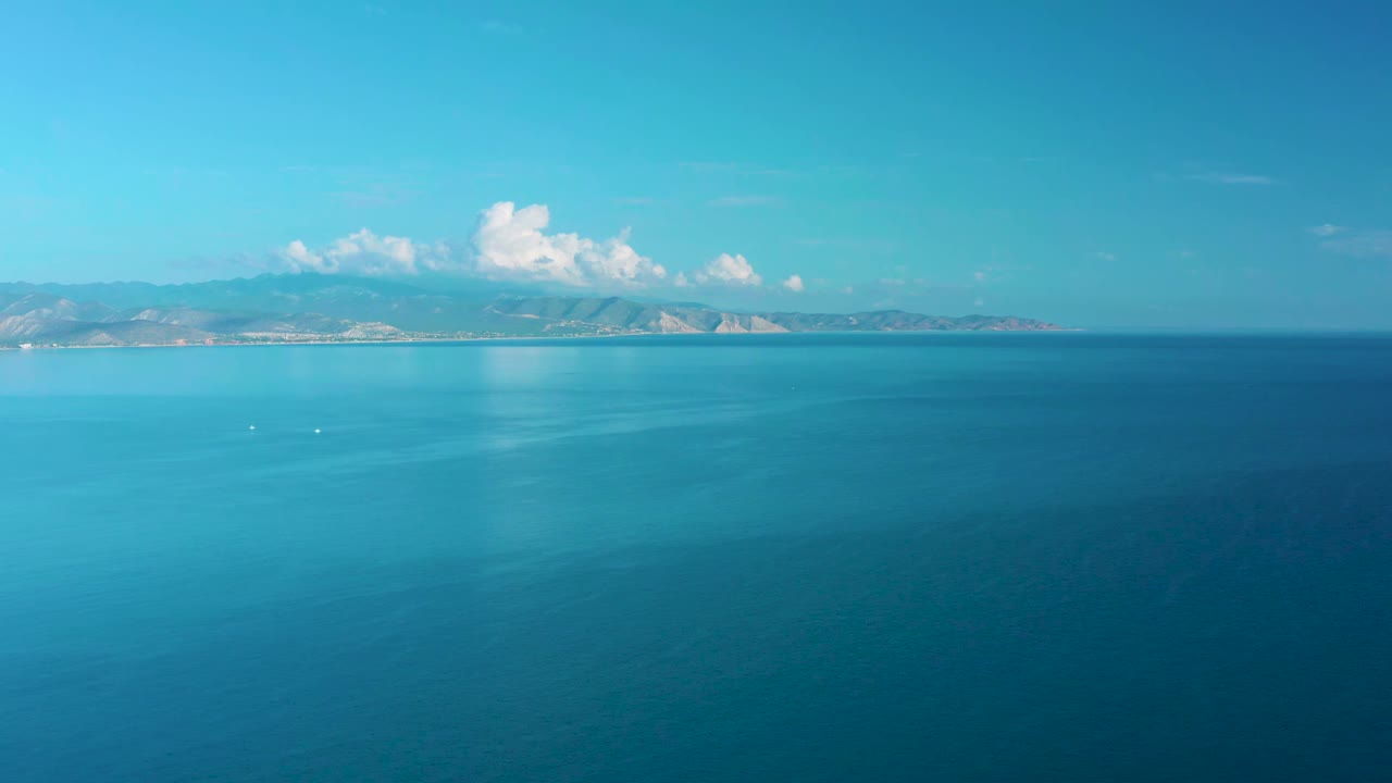 Turquoise water fills the foreground beneath a straight horizon, with the La Ribera coastline and bright cloud bank in the distance, no visible people.