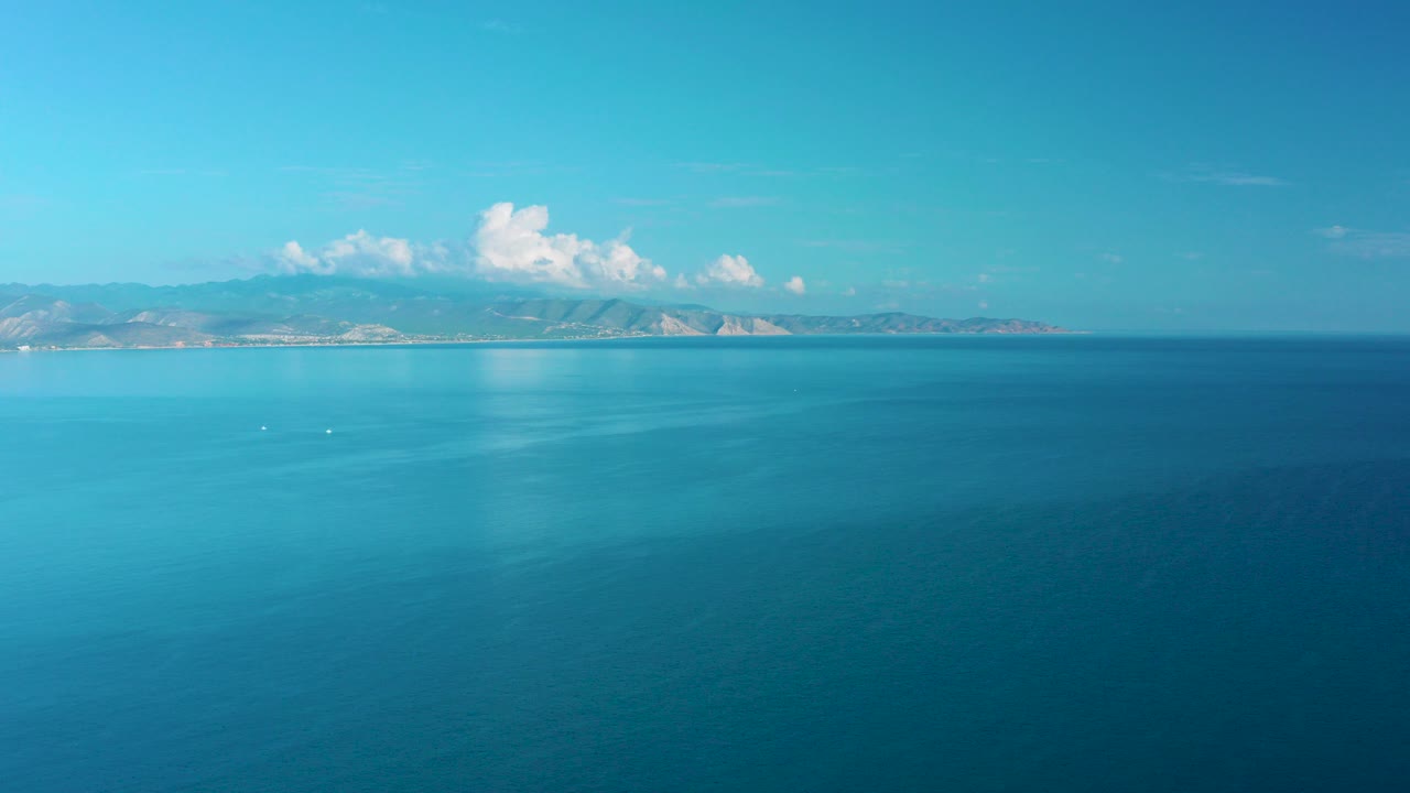 Teal ocean with distant hilly coastline under blue sky.