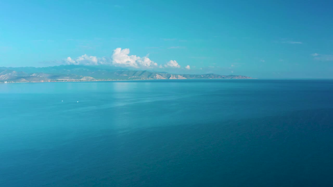 High aerial frame over blue-green water in La Ribera, with low coastal hills across the background, cloud reflections on the sea, and no visible people.
