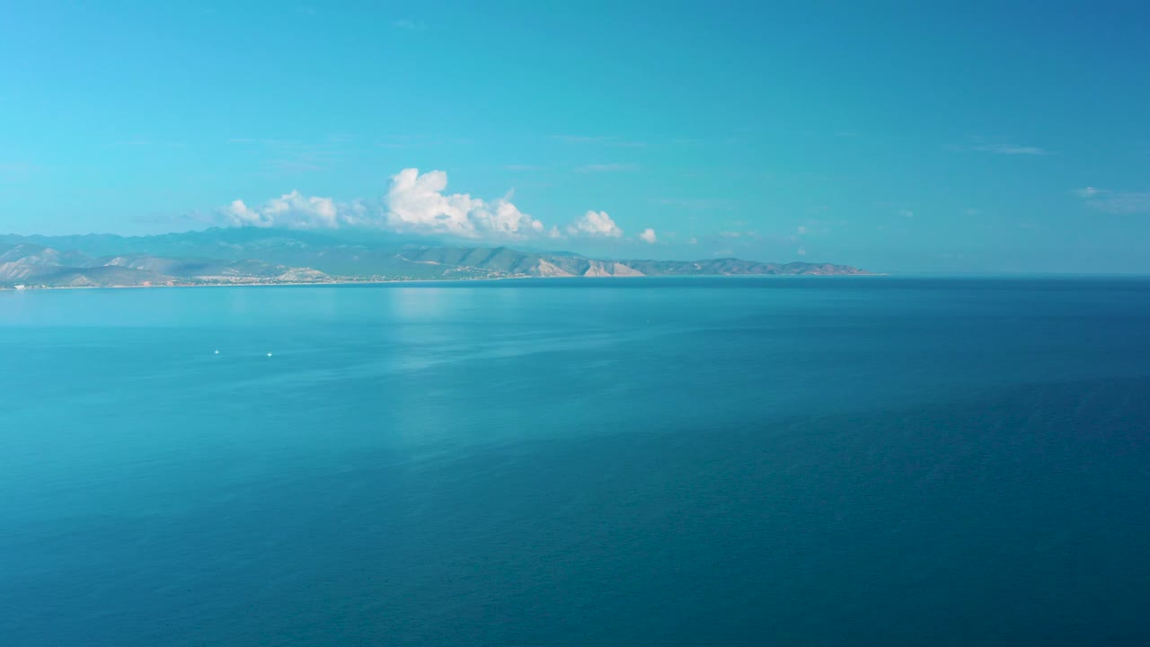 Aerial ocean view in La Ribera showing a darker blue foreground, lighter teal water near the horizon, distant hills, scattered white clouds, and no visible people.