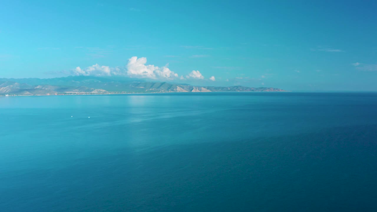 Mid-clip aerial frame near La Ribera with teal water filling the frame, low mountains along the horizon, bright white clouds above them, and no visible people.