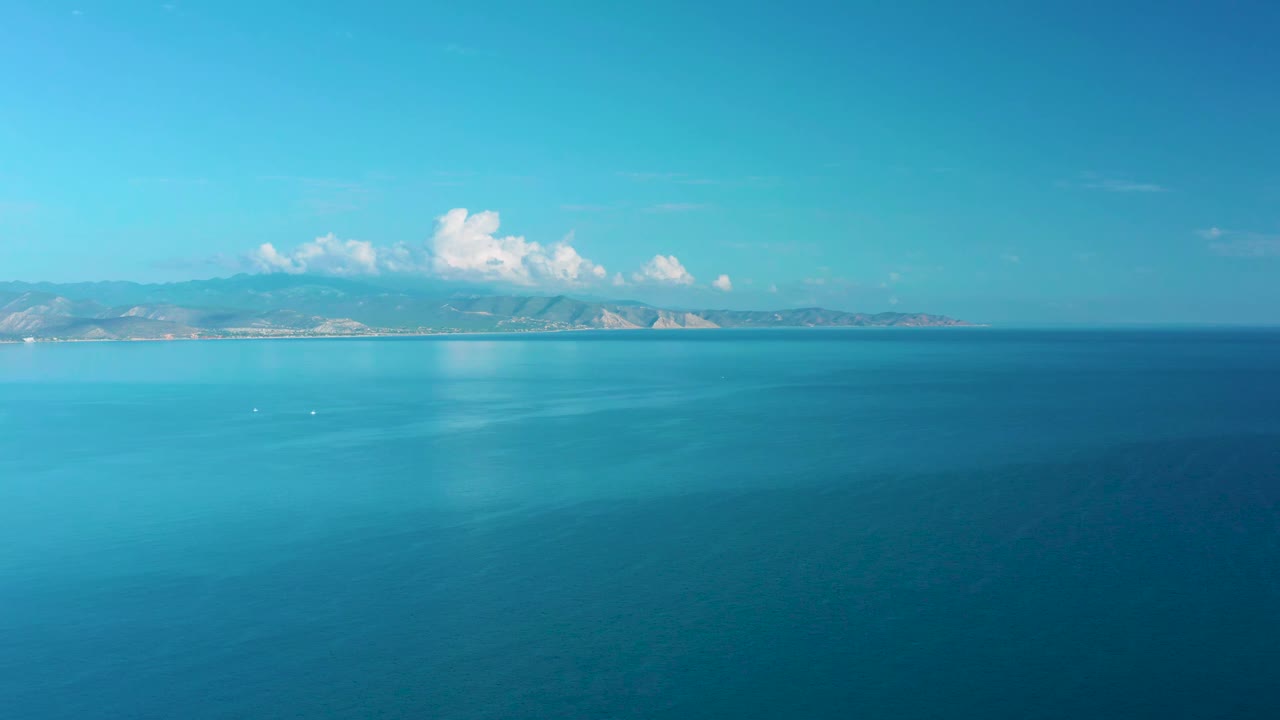 A wide aerial view shows blue-green ocean water, a thin distant coastline, a cloud cluster above the mountains, and no visible people in La Ribera, Baja California Sur.