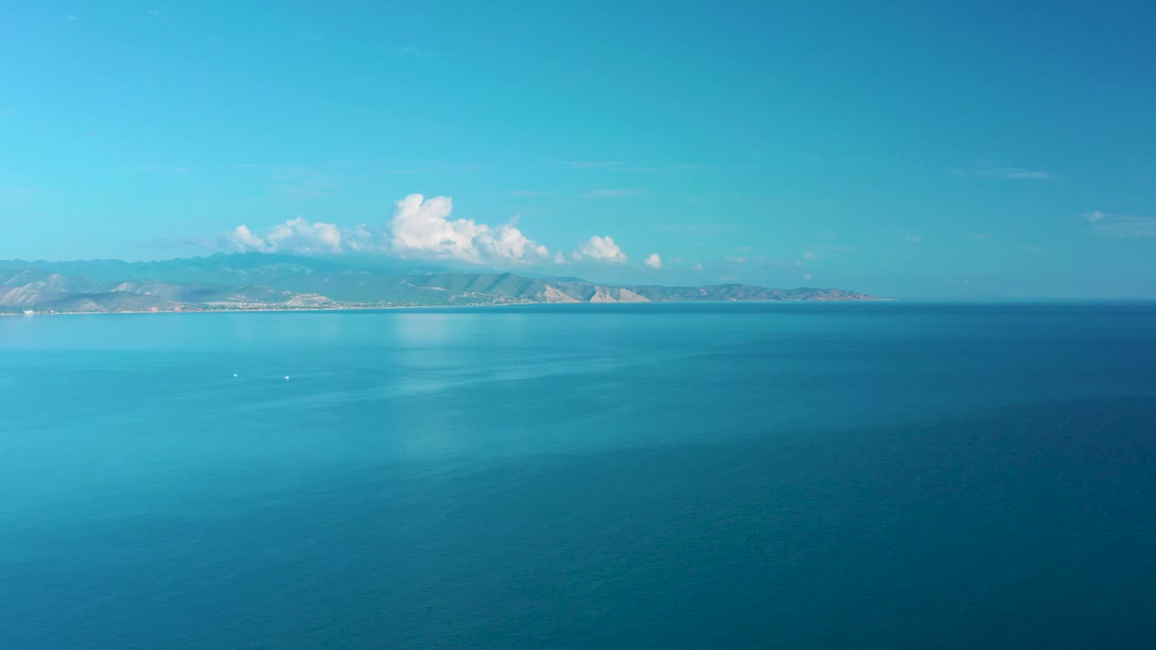 A broad stretch of calm ocean shows lighter blue patches on the water surface, distant mountains beneath white clouds, and no visible people in frame.
