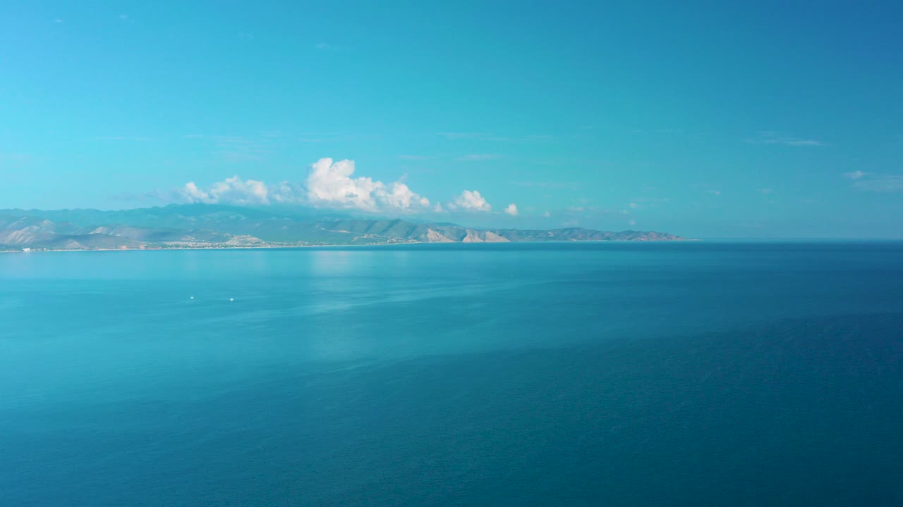 Wide aerial view over blue ocean toward the La Ribera coastline in Baja California Sur, with low mountains, white clouds, and no visible people.