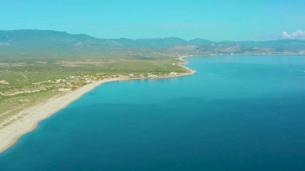 Wide aerial frame of La Ribera coastline with blue-green water occupying the right side, a pale beach curving through the left side, low buildings inland, hazy mountains, and no visible people.