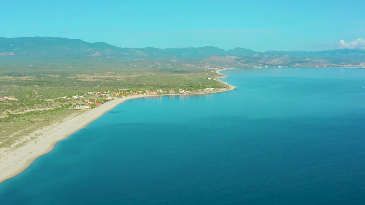 Wide aerial frame over La Ribera, Baja California Sur, showing shoreline homes near the beach, blue water filling the right side, mountains beyond, and no visible people.