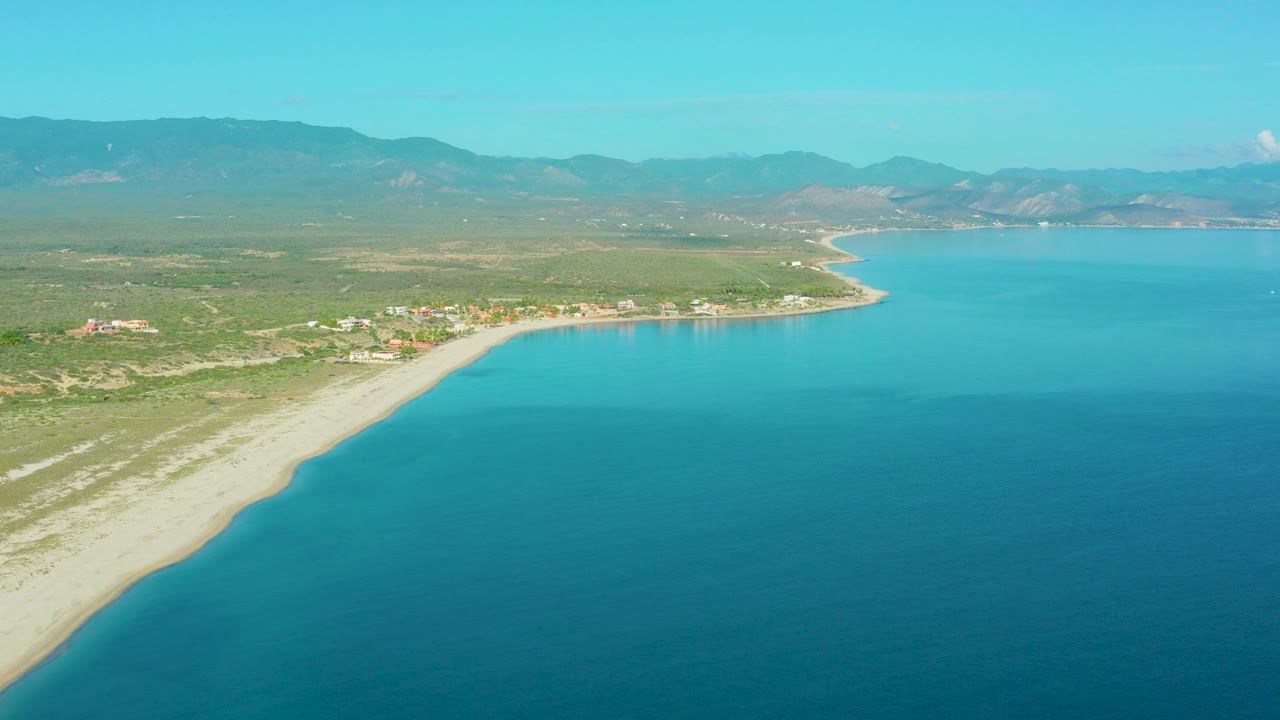 Aerial frame over La Ribera, Baja California Sur with turquoise water on the right, pale beach curving along coastal scrub, distant mountains, and no visible people.