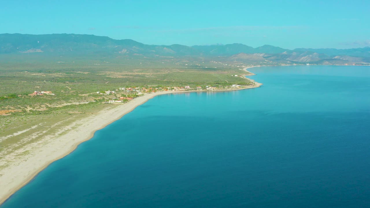 Aerial beach curve with turquoise water, inland scrub, buildings, and mountain backdrop.
