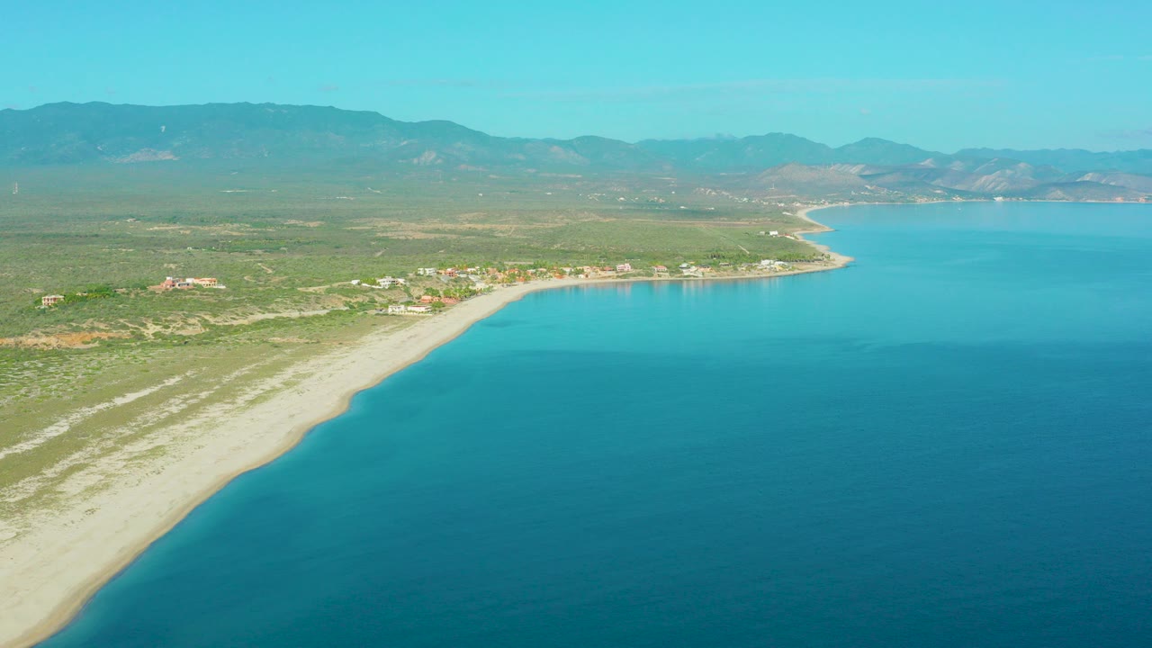 Aerial frame over La Ribera, Baja California Sur, showing blue seawater beside a pale sandy beach, low vegetation, inland buildings, and distant mountains, with no visible people.