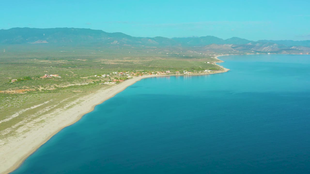 Aerial view in La Ribera with turquoise water filling the right side, a diagonal sandy shoreline, scrub-covered land, small inland structures, and a mountain range beyond, with no visible people.