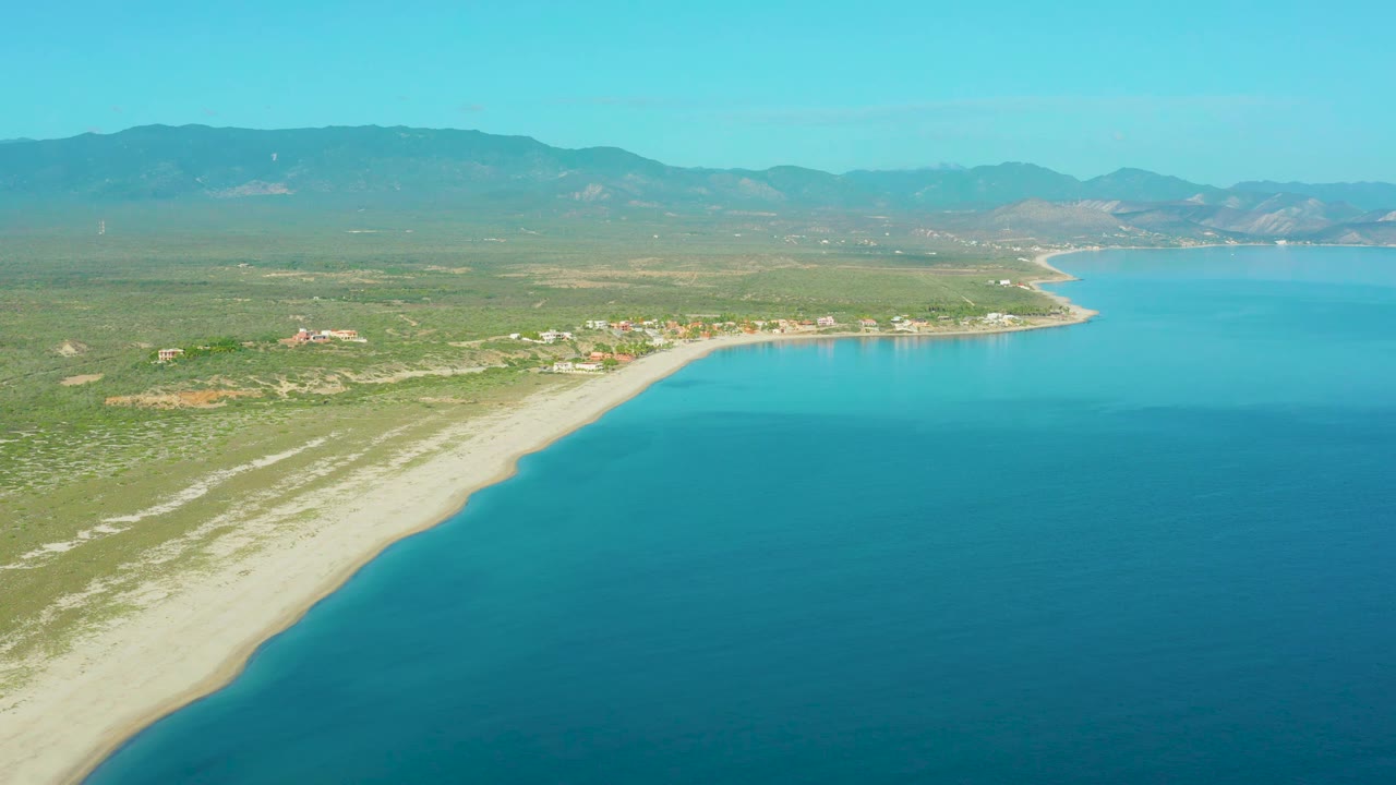 Aerial view over La Ribera showing the shoreline running diagonally from the lower left toward small coastal buildings, with calm blue water to the right and no visible people.