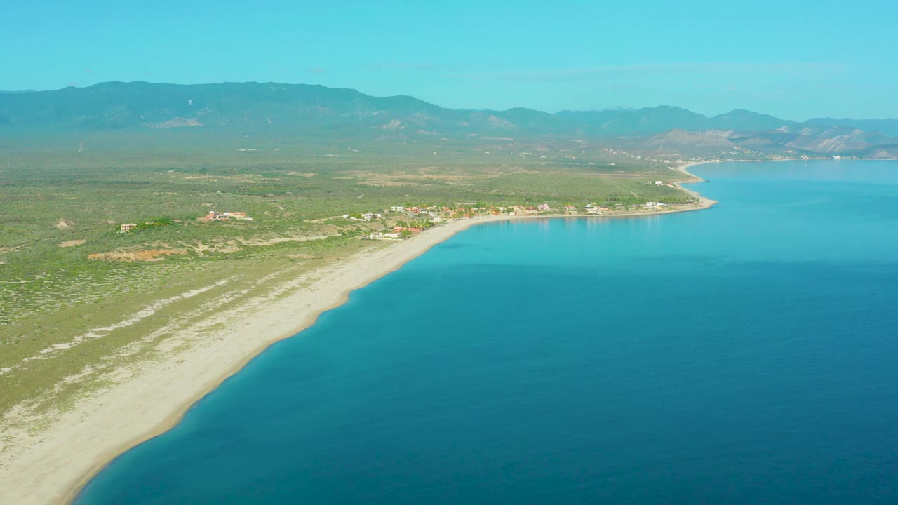 Mid-clip aerial view over La Ribera, with turquoise-blue ocean occupying the right foreground, pale beach curving left, low vegetation inland, distant hills, and no visible people.