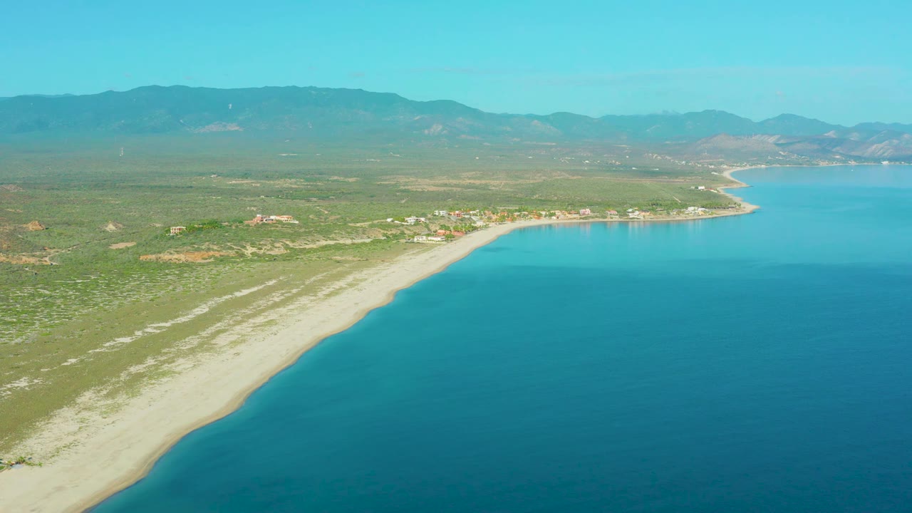 La Ribera coastline seen from above with the beach running diagonally across the frame, clear blue water on the right, sparse buildings inland, mountains beyond, and no visible people.