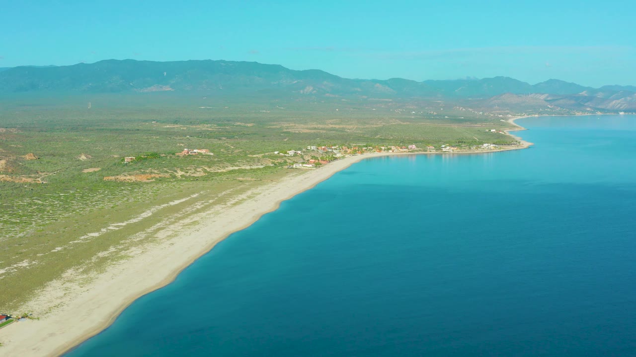 A wide La Ribera aerial frame shows more open water on the right, the pale beach tapering along the left edge, inland vegetation and buildings, a mountain horizon, and no visible people.