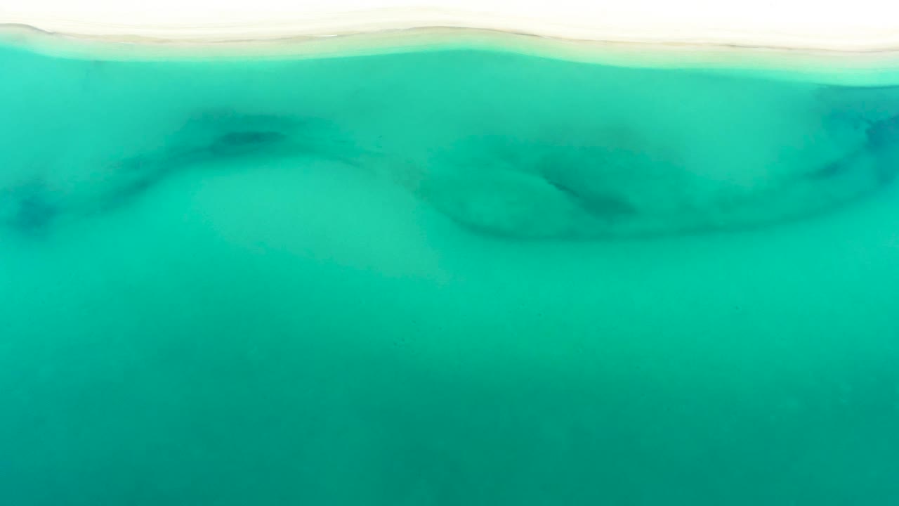 Aerial frame over La Ribera shoreline water, showing blue-green shallows, a narrow pale beach band at the top, and dark rock forms under the surface with no visible people.