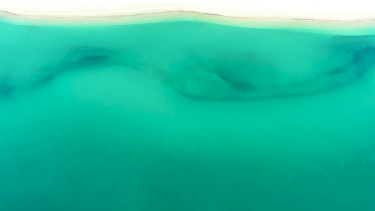 Top-down view at La Ribera, Mexico, with bright turquoise water, a thin sand line across the upper frame, and cloudy dark rock texture beneath the surface, no visible people.