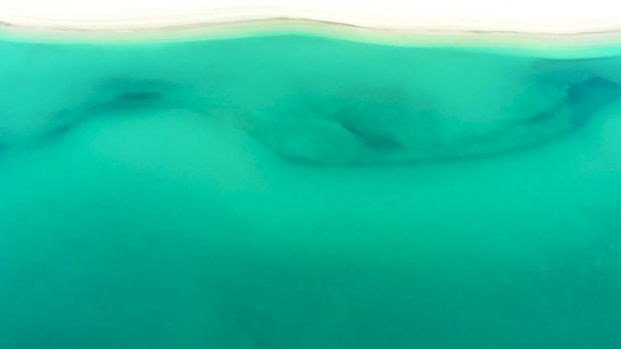 Overhead La Ribera water detail with the pale sandy shoreline along the top, broad turquoise shallows through the center, and darker submerged formations faintly visible, no visible people.
