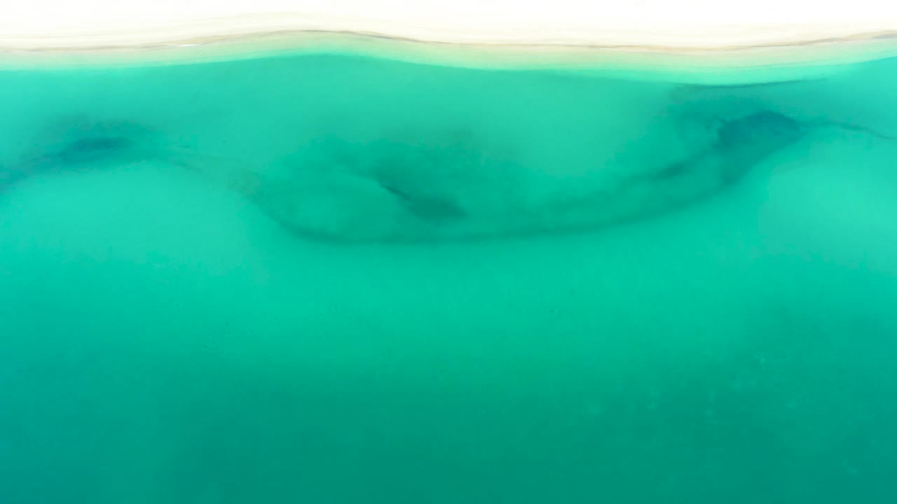Clear blue-green saltwater borders the pale beach edge in La Ribera, with a broad darker underwater patch near the upper center and no visible people.