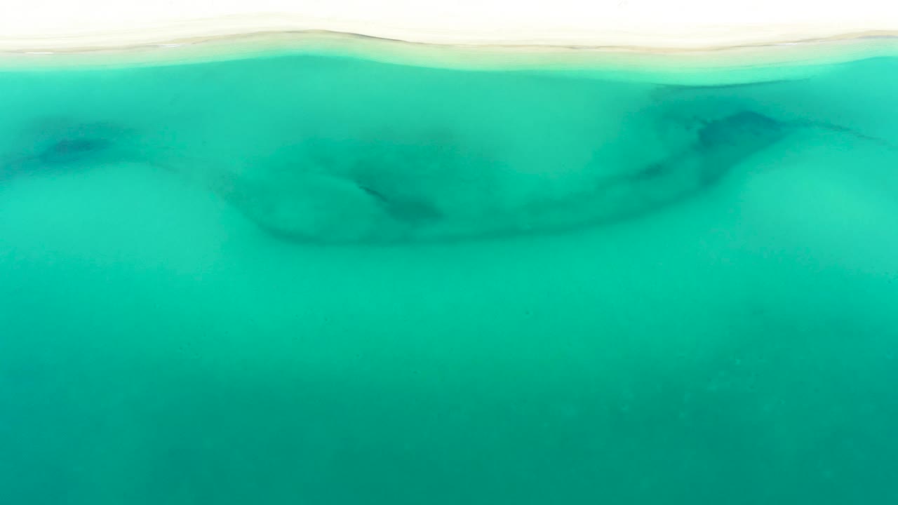 Aerial-style view of La Ribera shoreline water, where pale sand meets turquoise sea and darker submerged forms sit just below the surface, no visible people.