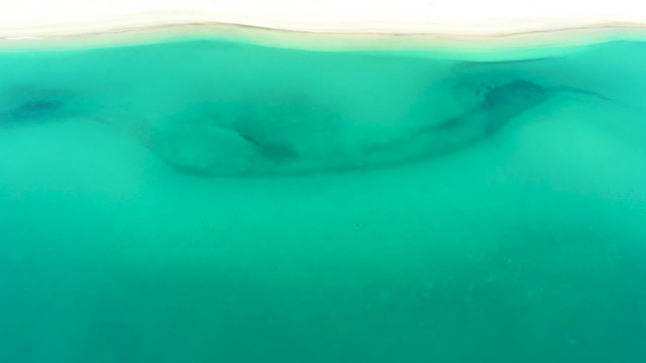 The La Ribera shoreline remains near the top of frame, with bright turquoise water below it, soft underwater shadows, and no visible people.