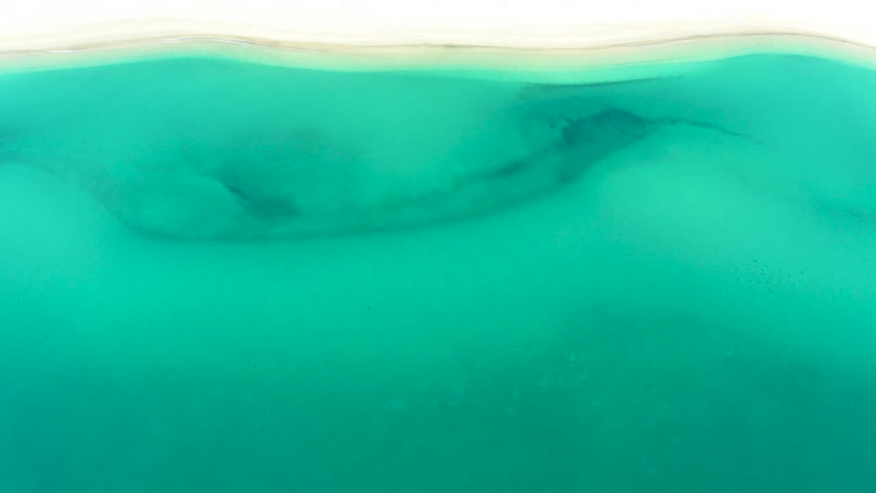 Overhead view of turquoise water meeting a pale sandy edge in La Ribera, Baja California Sur, with darker underwater shapes and no visible people.