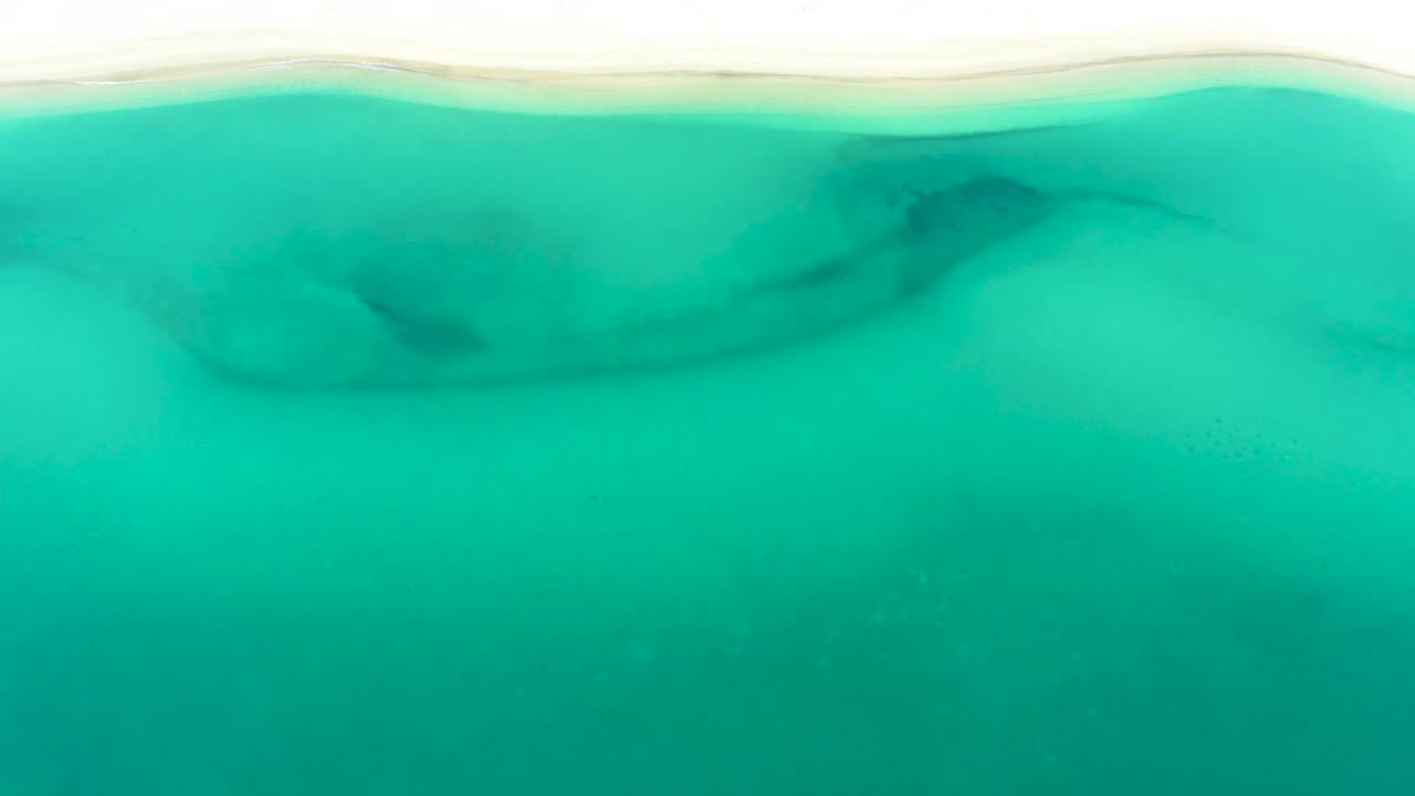 Clear teal shallows run below a narrow bright sand strip in La Ribera, with soft darker sandbar forms under the water and no visible people.
