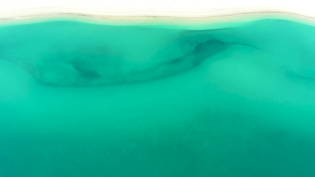 Aerial view of turquoise shoreline water in La Ribera, Baja California Sur, where darker underwater patches sit below the pale beach edge and no visible people appear.