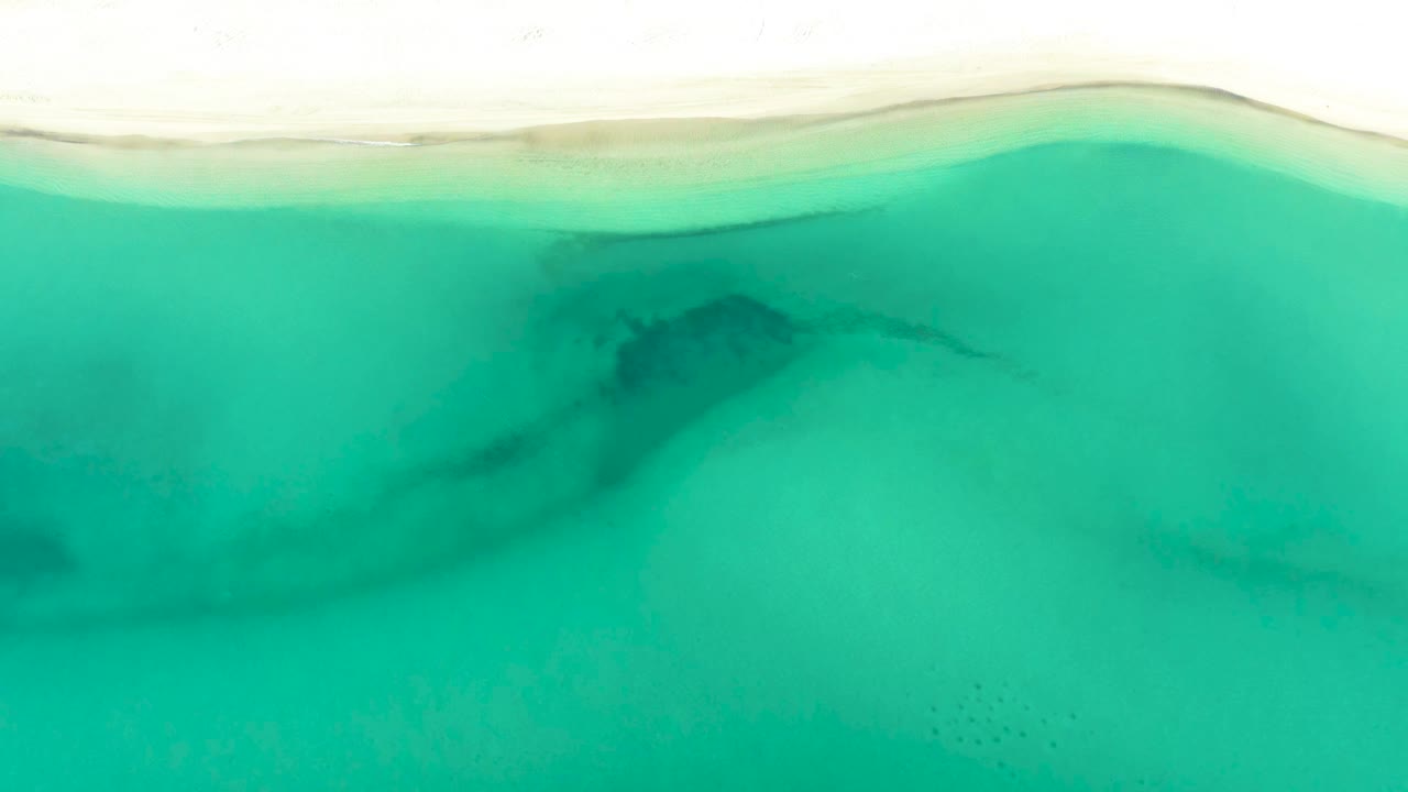 Aerial beach edge above turquoise water with a darker patch near center.