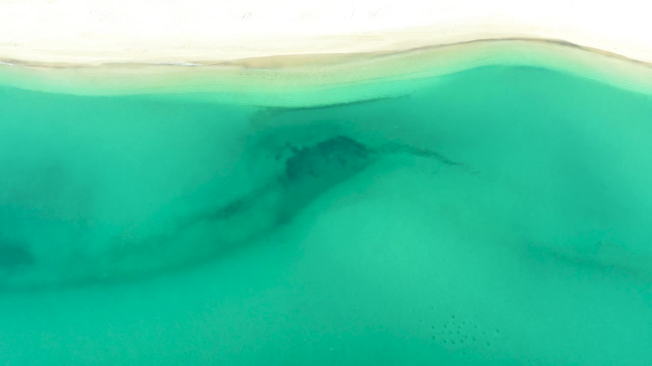 High drone view over La Ribera with a pale beach strip at the top, translucent turquoise water filling the frame, a shadowy underwater patch to the left of center, and tiny distant people near the lower water.