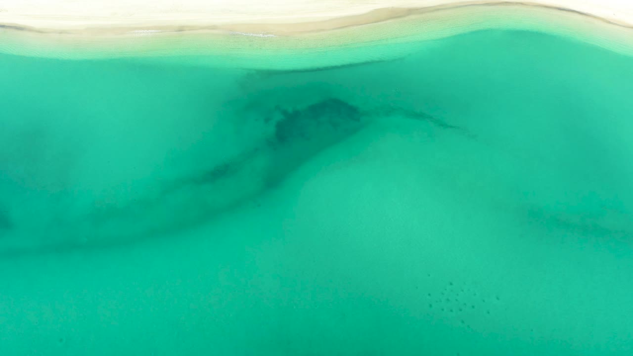 Aerial-looking frame of turquoise-green water below a pale beach edge in La Ribera, Baja California Sur, with no visible people.