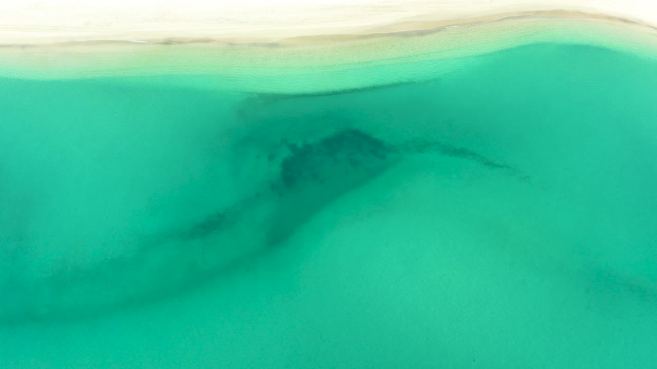 Overhead view of turquoise water and pale sand in La Ribera, Baja California Sur, with a dark underwater patch near the left-center and no visible people.