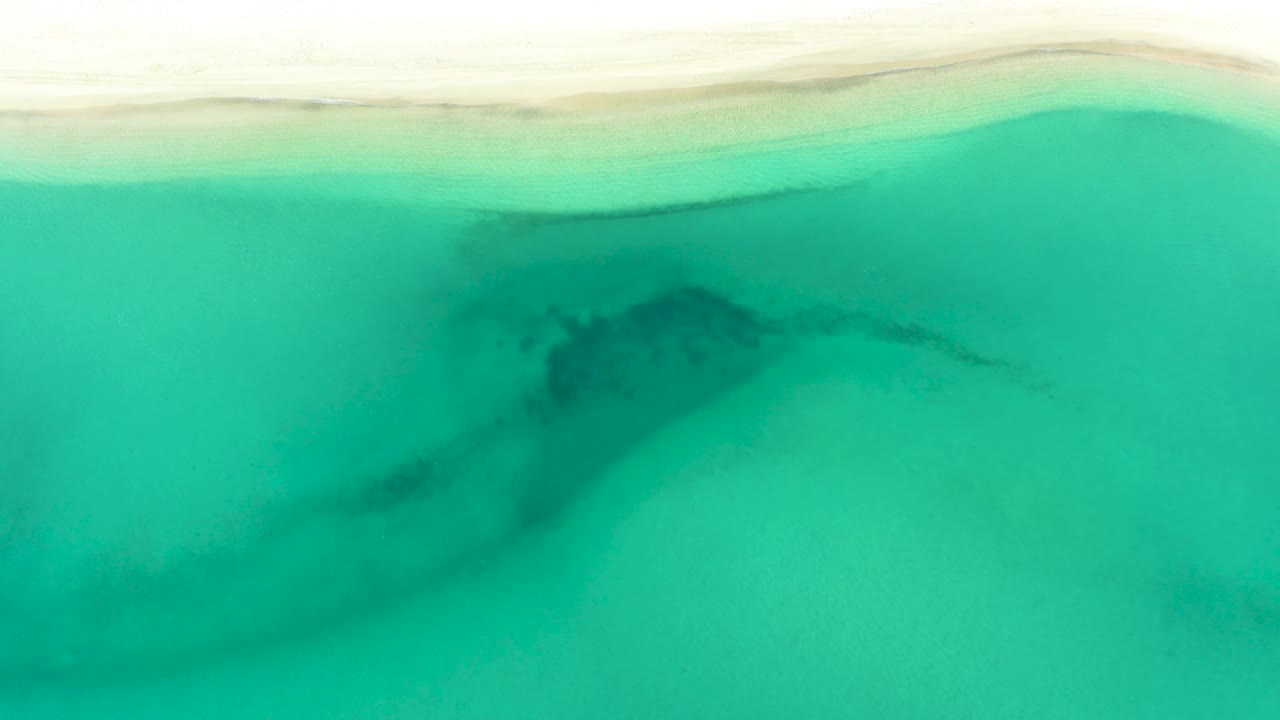 Overhead coastal view in La Ribera showing a bright turquoise water field, a thin pale beach strip at the top, darker submerged markings, and no visible people.